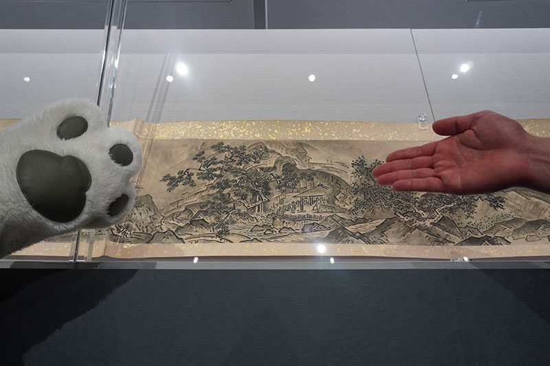 雪舟筆四季山水図巻模本（狩野探幽筆、京都国立博物館所蔵）を中心に、向かって左にトラりんのもふもふとした手、右に福士研究員の手
