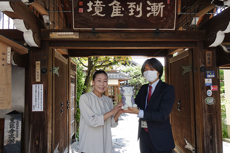 瑞泉寺山門前にて、ミニトラりんを抱える山内研究員と羽田研究員