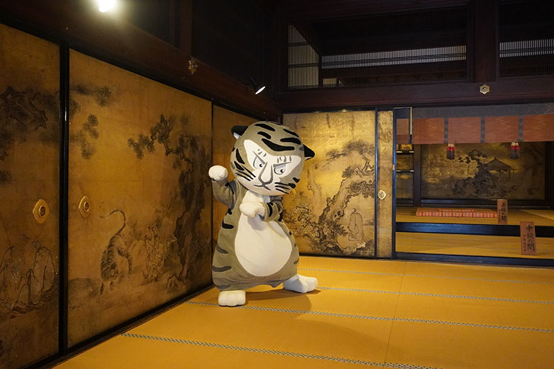 仙人の間にて、虎が描かれた障壁画の前でポーズをとるトラりん