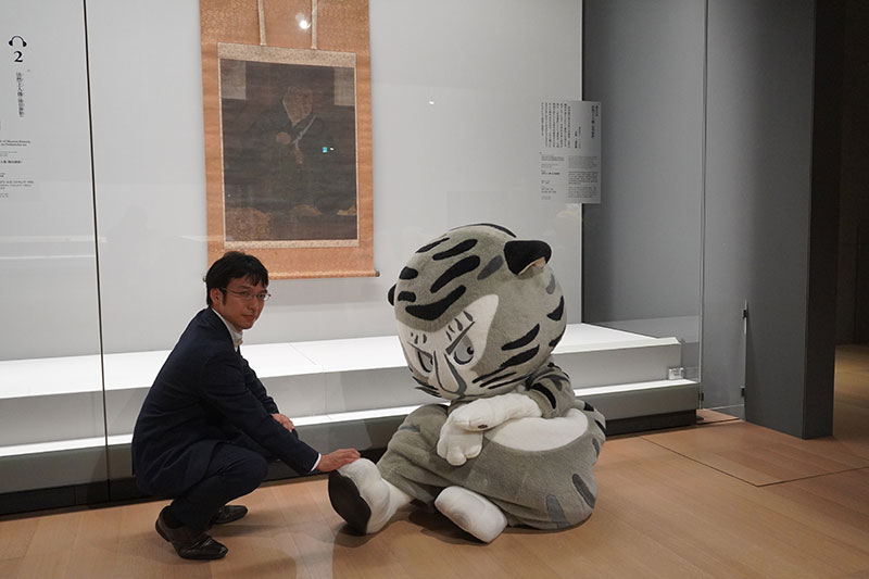 肖像をまねてあんよを出すトラりんと、念ではなく手で押して、あんよを引っ込めようとさせる井並研究員