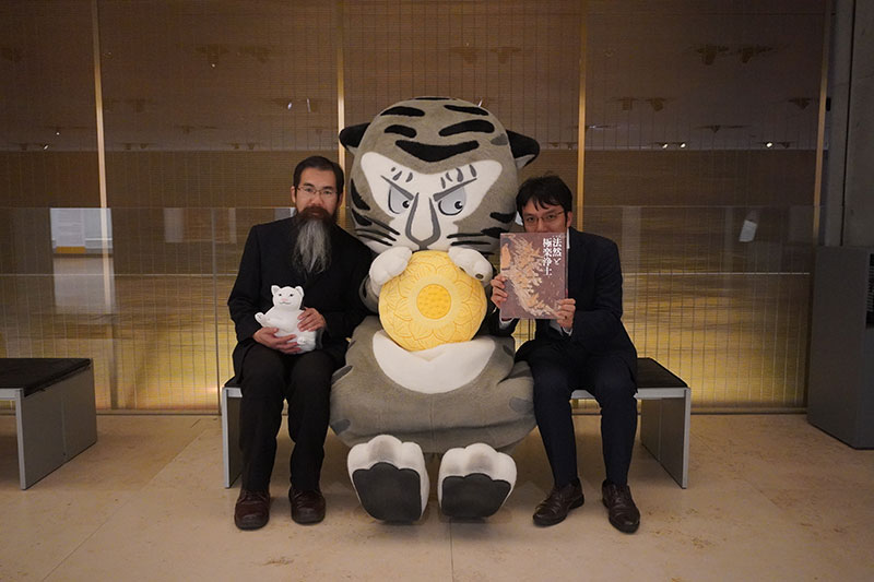 ハチワレネコぬいぐるみを持つ大原研究員、蓮台クッションを持つトラりん、図録を持つ井並研究員
