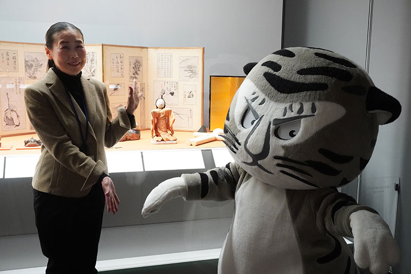 「這子」の展示ケース前、人形のポーズのまねて、手をだらりとするトラりん