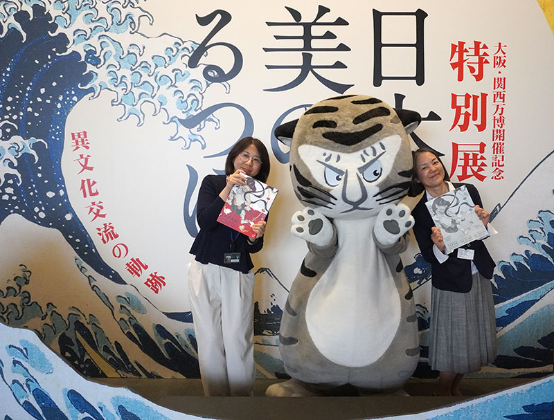 1Fの看板前、図録をもつ永島研究員、石田研究員