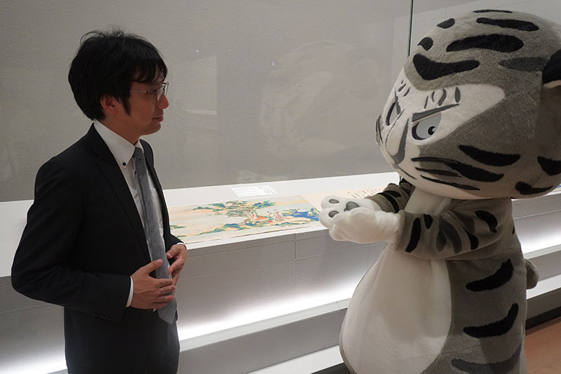 展示ケースの前で説明する井並研究員と乳粥をもつポーズのトラりん