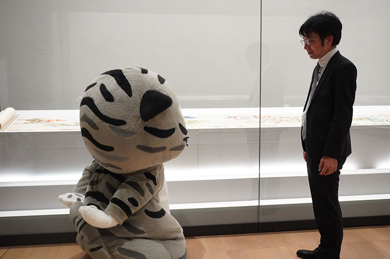 展示ケースの前で説明する井並研究員と背負うポーズのトラりん