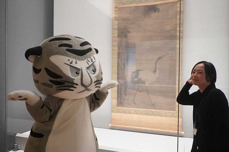 「観音猿鶴図」左幅の前で、耳を澄ませる森橋研究員と鶴の真似をするトラりん