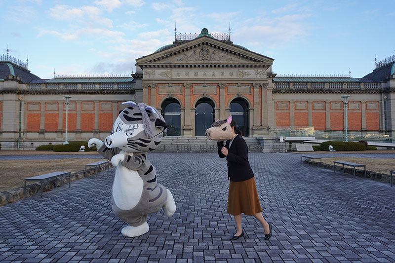 水谷研究員・トラりんが展示室に向けて歩を進めるポーズ
