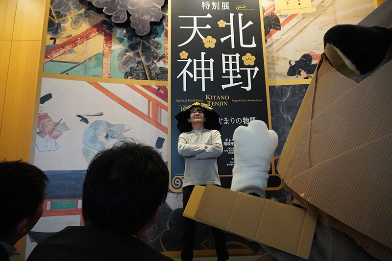 看板前に仁王立ちになり腕組みをする末兼研究員。崇め奉るようなポーズをとるトラたち