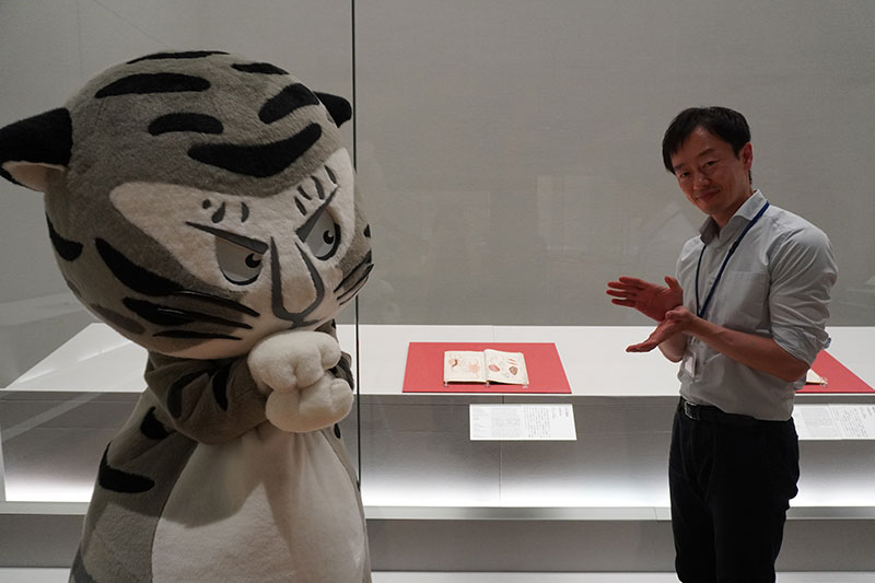 作品の前で、手を二枚貝に見立てて、貝が開くポーズをするトラりんと福士研究員