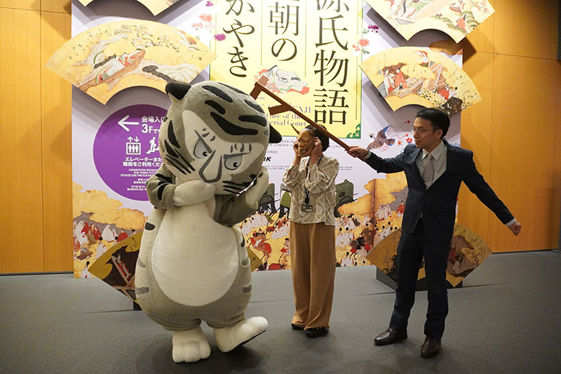 エントランスロビー看板前で、般若面をつけた山内研究員、トラりんを打杖で威嚇する井並研究員