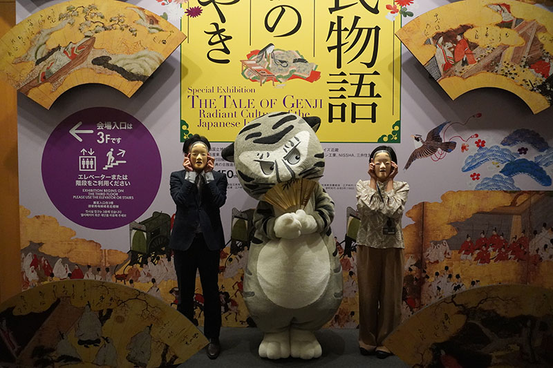 エントランスロビー看板前で能面をつけた研究員と扇をもつトラりん